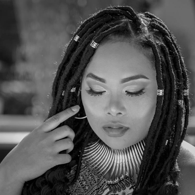 Black-and-white portrait of a woman with locs, looking downward with a hand beside her face.